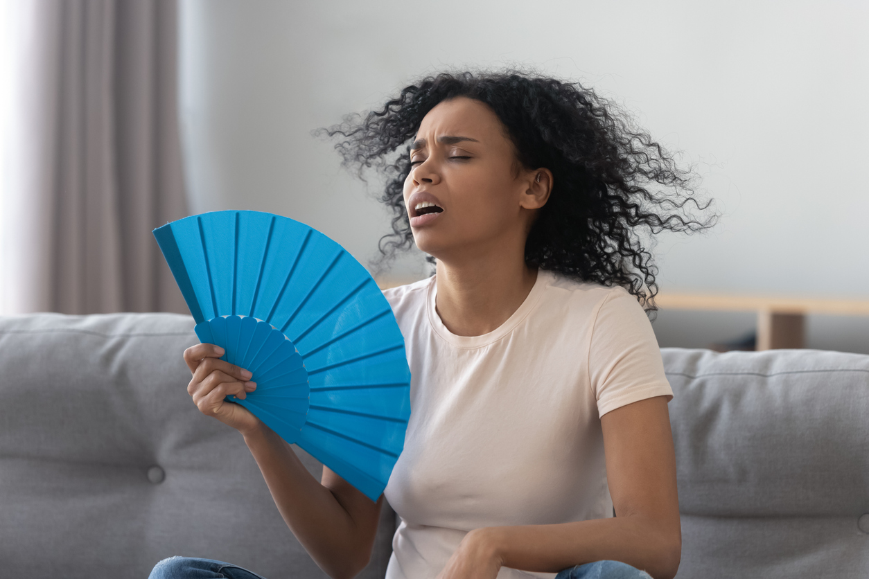 Overheated african young woman feeling hot waving fan annoyed with high temperature sit on sofa at home, stressed black girl sweating suffer from summer weather heat problem without air conditioner