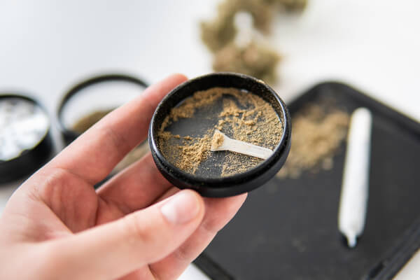 A hand holding up the Kief catcher of a grinder with a joint and rolling tray in the background