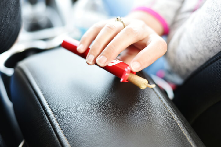 Mature woman passing joint to passenger in back, sticking out of red plastic container while driving.