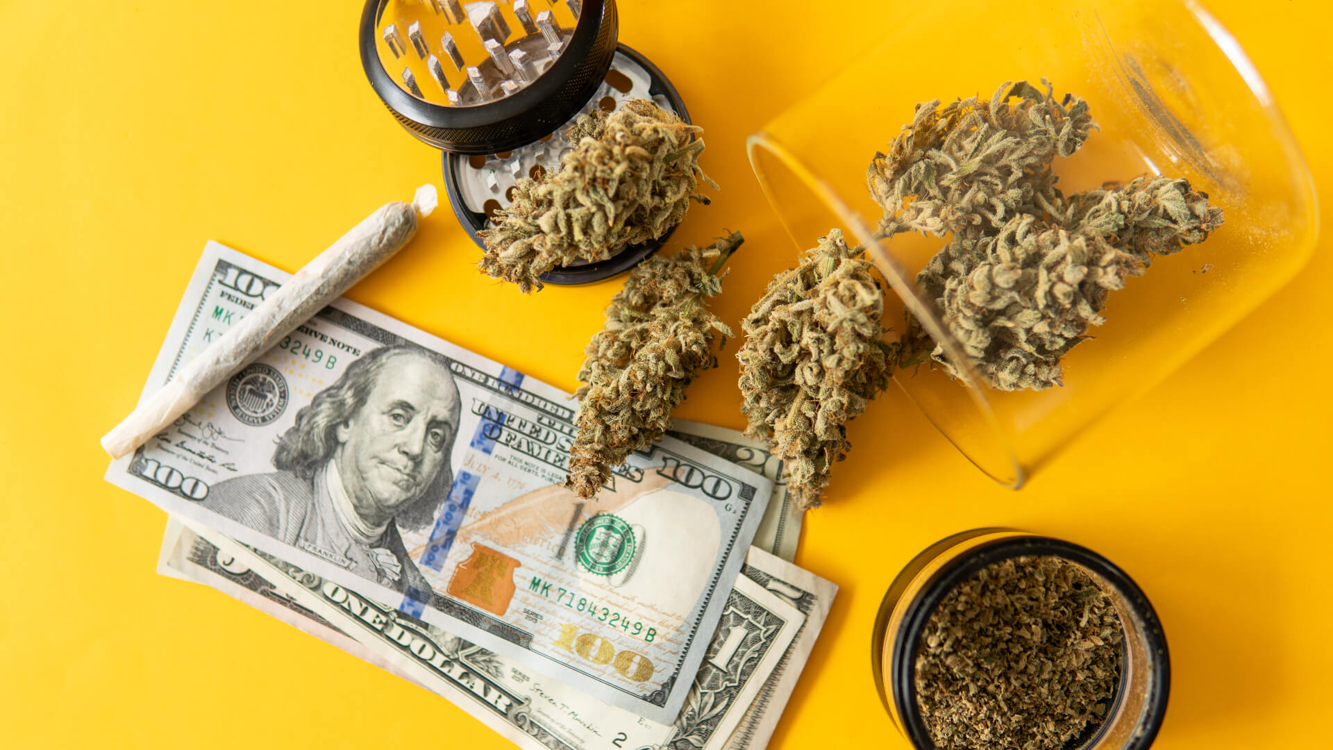 Yellow background with money, joints, a grinder, and green buds lying on the surface