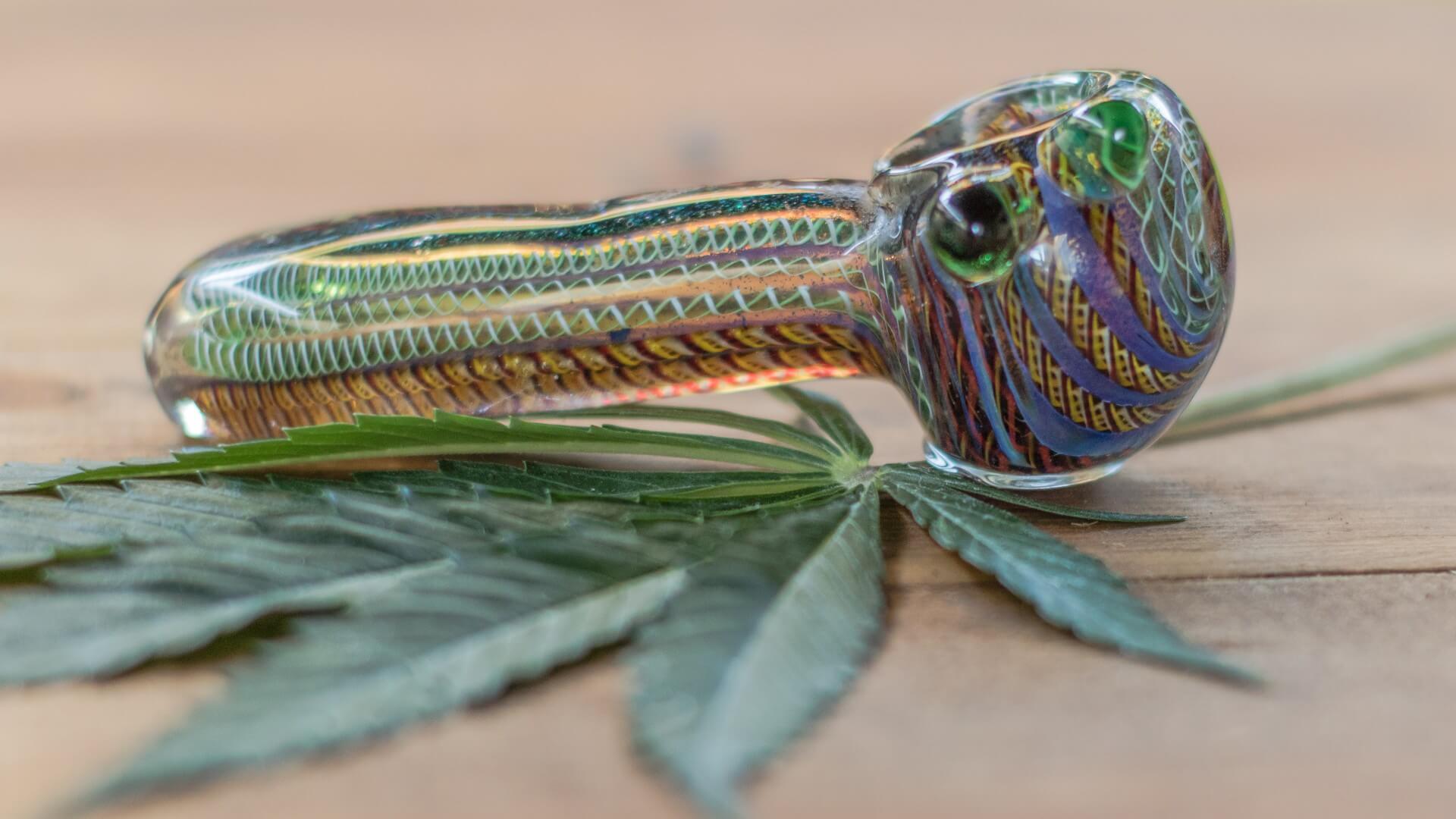 A glass blown cannabis pipe with swirling colors resting on top of a cannabis leaf.