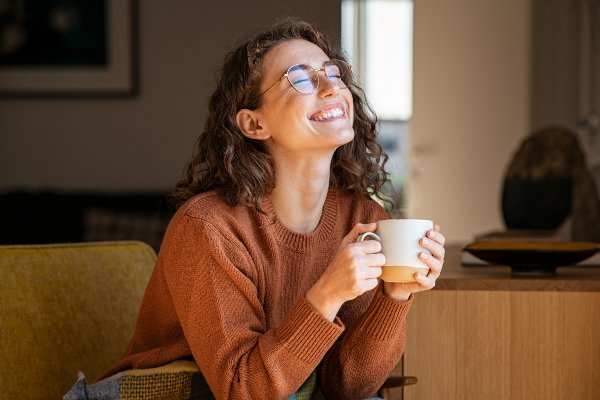 woman holding tan coffee cup and smiling with her eyes shut while wearing orange sweater