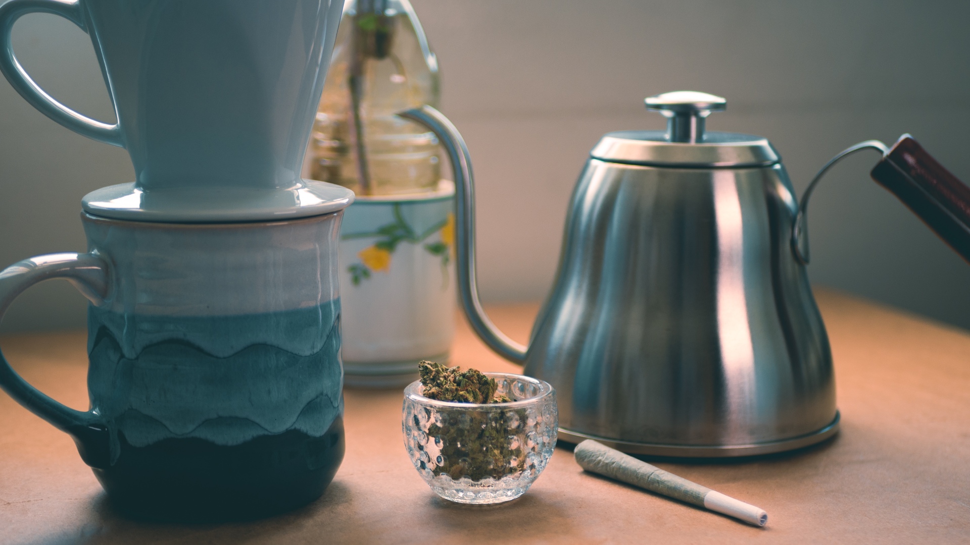 A blue ceramic pour over coffee cup sitting next to a joint and a metal kettle